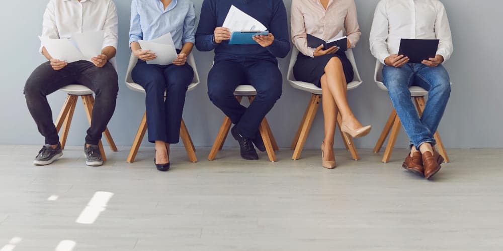 Cinco pessoas sentadas lado a lado em uma fileira de cadeiras esperando por uma entrevista de admissão