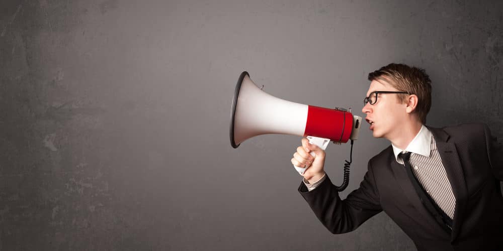 Homem vestindo terno preto e óculos de grau segura um megafone branco e vermelho em frente à boca