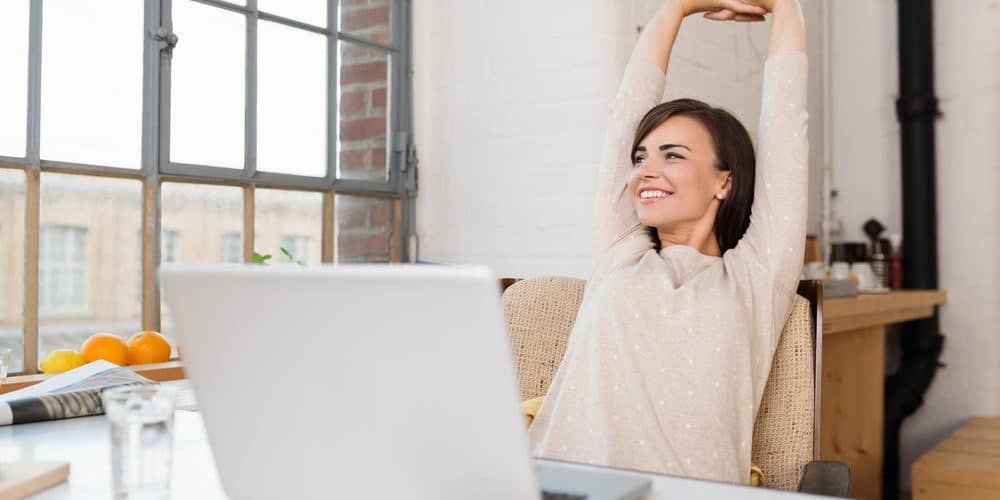 Mulher de cabelos escuros curtos sorri enquanto se espreguiça em frente á sua mesa de trabalho