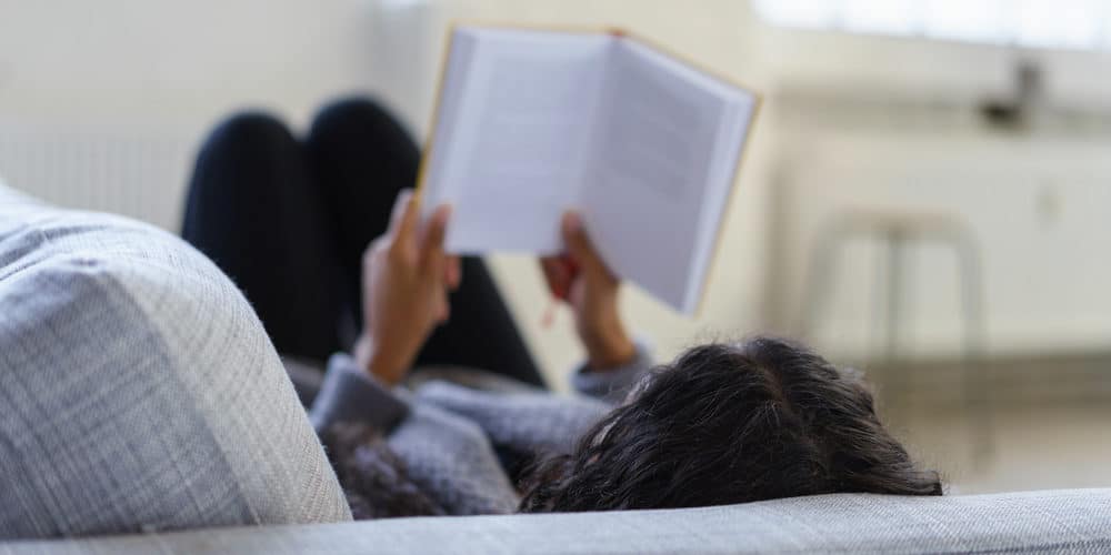 Mulher de cabelos castanhos está deitada no sofá de sua casa enquanto lê um livro em seu dia de descanso semanal remunerado