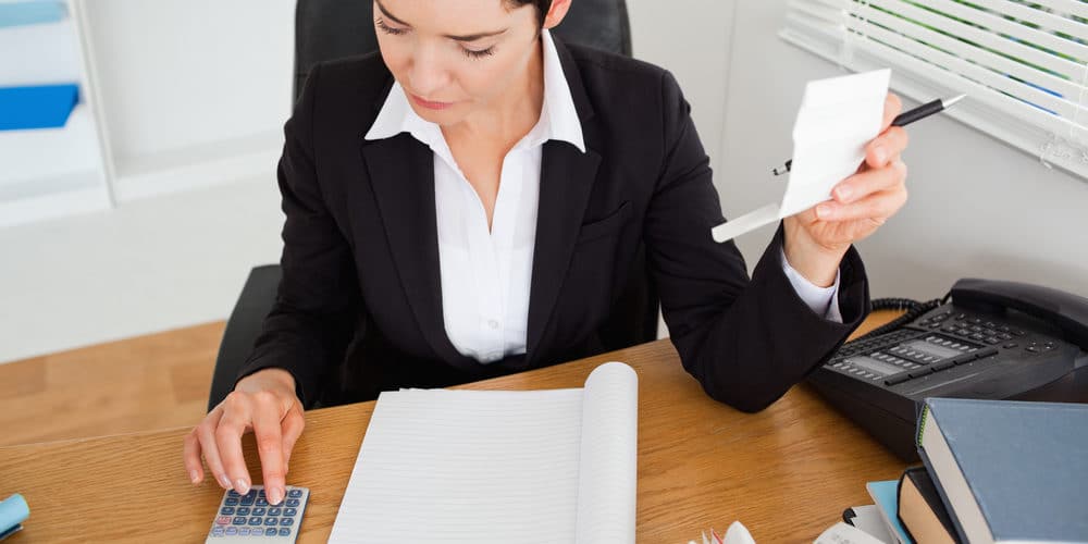 Mulher vestida com roupa social faz cálculos da folha de pagamento numa calculadora em sua mesa de escritório