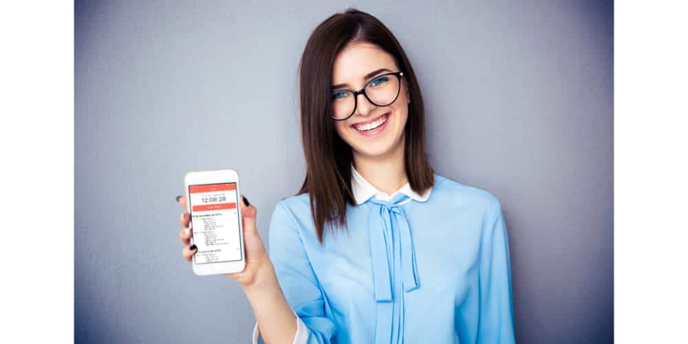 Mulher branca, de cabelos escuros, usando camisa azul claro e óculos de armação escura está sorrindo para a câmera e mostrando um smartphone com o aplicativo de controle de ponto da mywork aberto