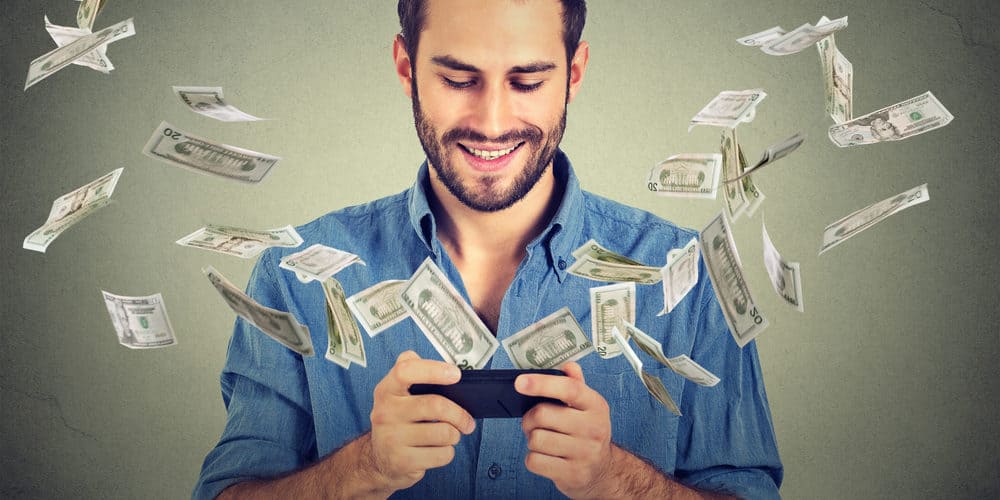 Homem de camisa azul olhando para a tela do celular enquanto cédulas de dinheiro caem ao redor dele