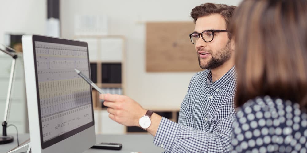 Homem de cabelos pretos e óculos de armação escura explica informação em tela de computador para sua colega que está sentada ao lado