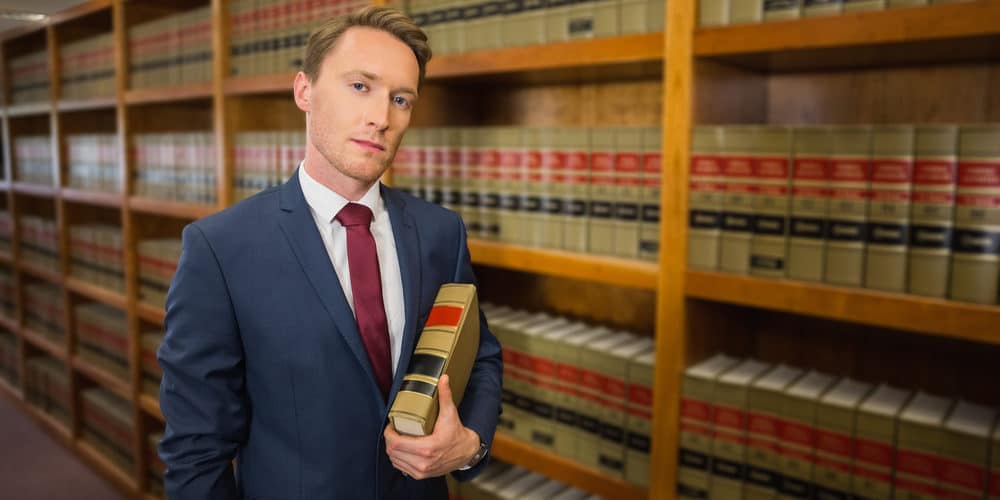 Homem branco loiro vestido terno social e carregando um livro posa para a foto em frente à estante de biblioteca