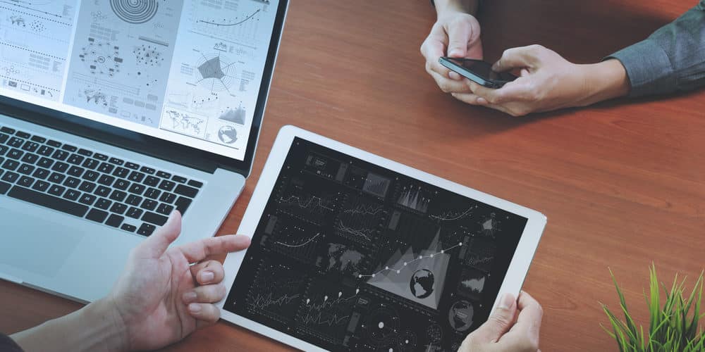 Pessoa mexendo em tablet branco sobre mesa de escritório com um laptop aberto sobre o tampo da mesa ao lado