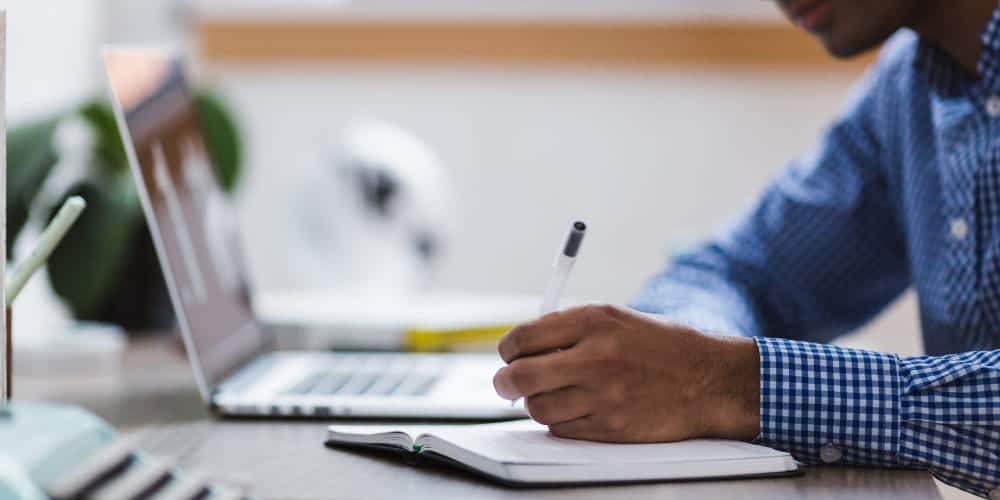 Homem trabalhando em mesa de escritório enquanto anota informações num caderno pequeno e checa informações em seu laptop