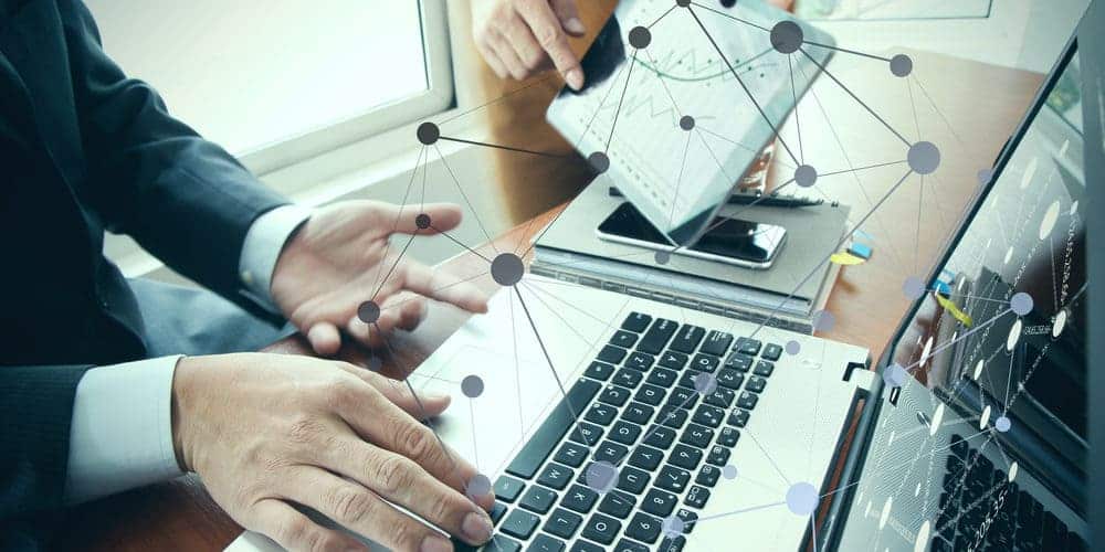 Imagem focada na mesa de um trabalhador que está mexendo em um computador prateado com teclas pretas