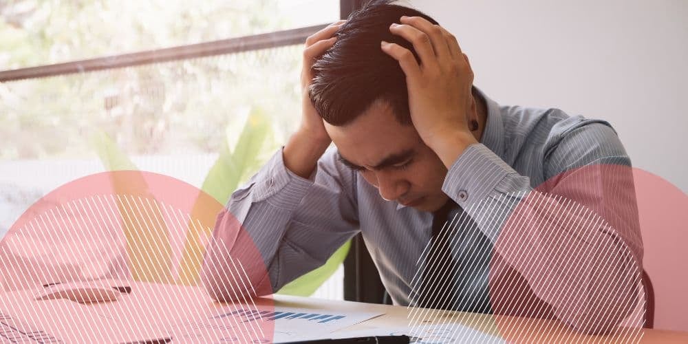 Homem branco, com feições preocupadas, vestindo roupa social, no ambiente de trabalho sentado à mesa de trabalho com as mãos apoiadas na cabeça