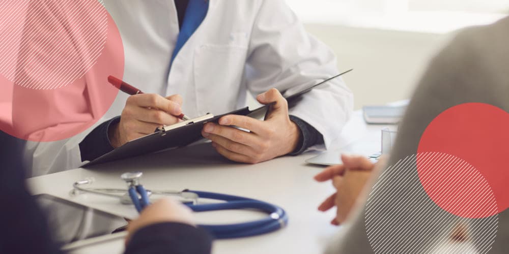 Médico vestindo jaleco branco assinando um atestado sobre prancheta enquanto fala com dois pacientes sentados do outro lado da mesa