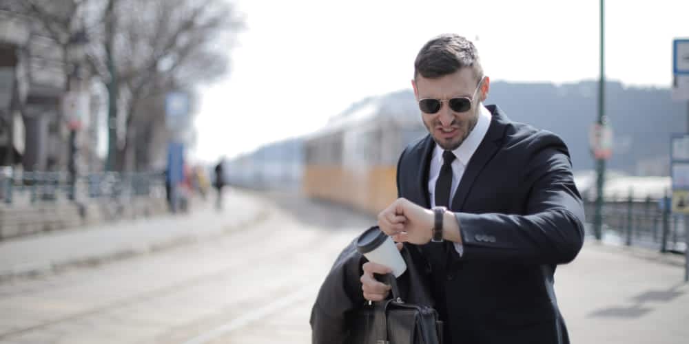 Homem vestindo terno e óculos escuros checa preocupadamente as horas em seu relógio de pulso enquanto espera o trem