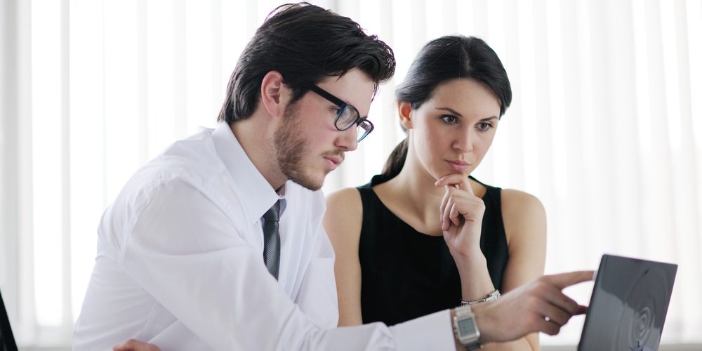 Homem de camisa, gravata e óculos e mulher de vestido regata e cabelos presos trabalham juntos e estão sentados em frente a uma mesa de escritório enquanto conversam sobre trabalho