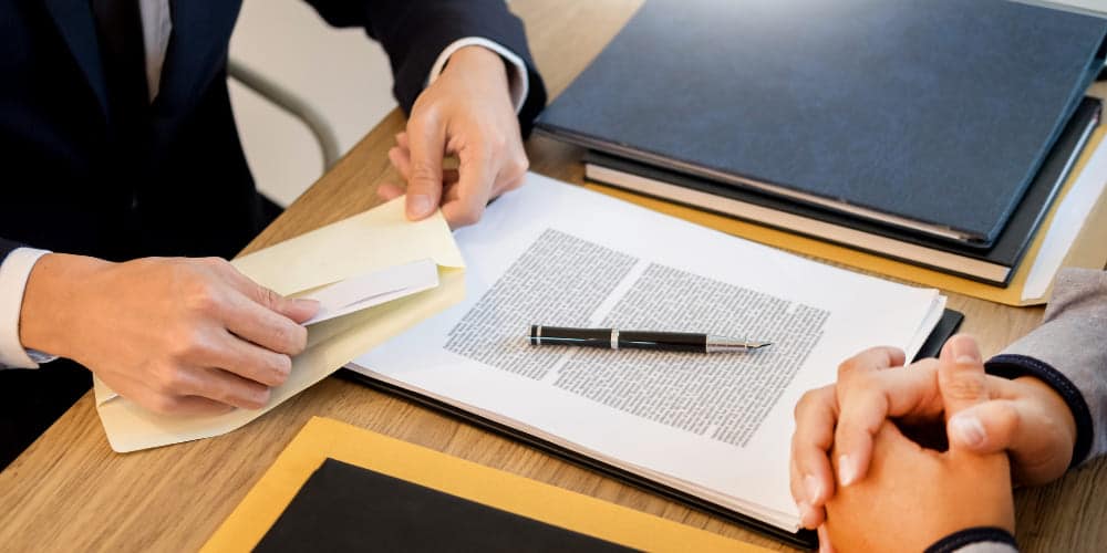 Documento com caneta preta em cima de uma mesa de trabalho perto de duas pessoas que estão sentadas em frente à mesa