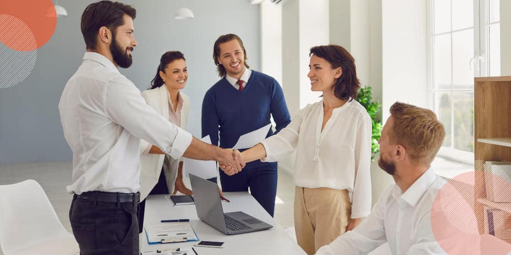 Grupo de cinco trabalhadores em sala de escritório observando enquanto dois deles, um homem e uma mulher, ambos vestindo camisas brancas, apertam as mãos.
