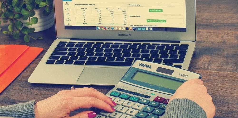 Mãos femininas com unhas pintadas de vermelho mexendo em uma calculadora e em um computador com planilhas abertas