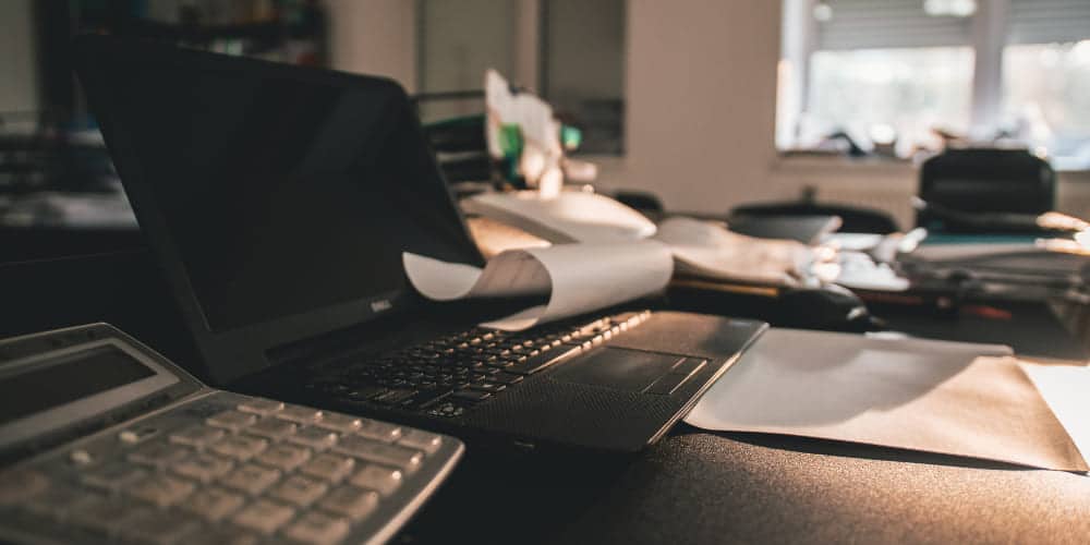Escritório de trabalho à meia luz com mesa de tampo escuro sobre a qual repousam documentos, cadernos, calculadoras e outros itens de papelaria