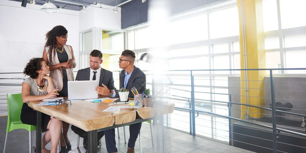 Grupo de colegas de trabalho reunidos em uma mesa de madeira dentro de uma sala de reunião de um escritório enquanto falam sobre projetos de trabalho em frente a um computador