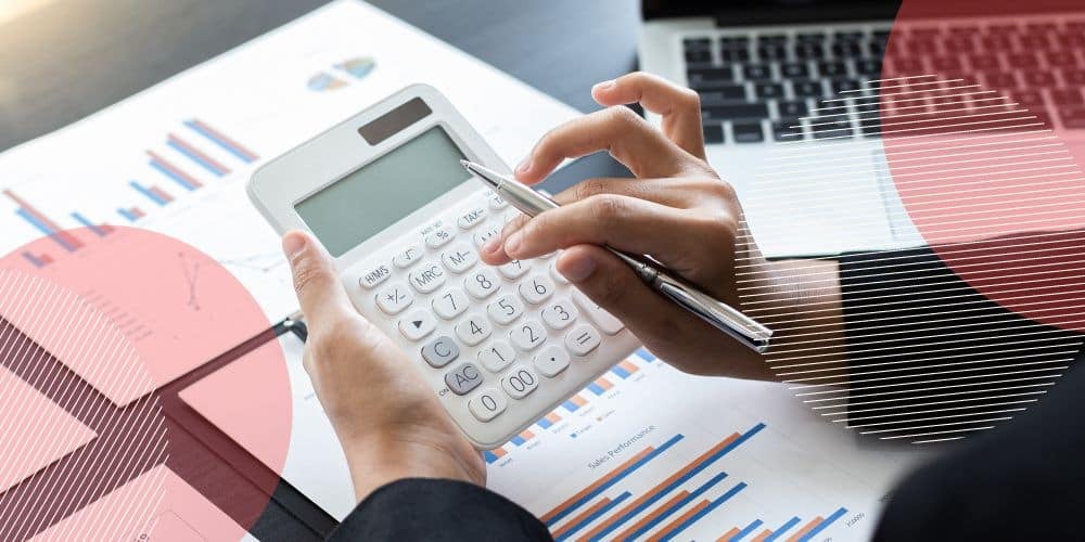 Mulher segurando uma caneta prateada na mão direita enquanto faz contas usando uma calculadora branca sobre uma mesa com documentos e um computador