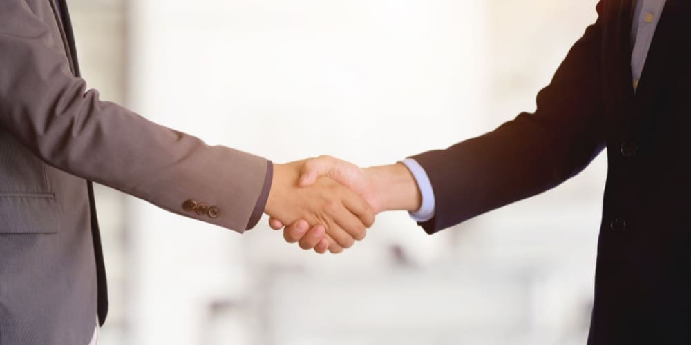 Duas pessoas vestidas com roupas sociais de trabalho apertam as mãos em frente à câmera 