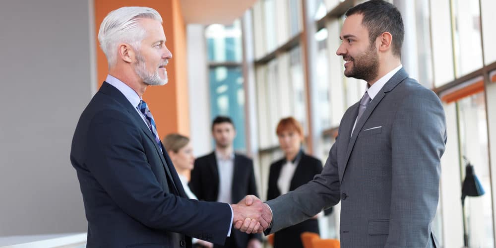 Homem de cabelos brancos vestido com terno social aperta mão de homem de cabelos castanhos também vestido de terno social