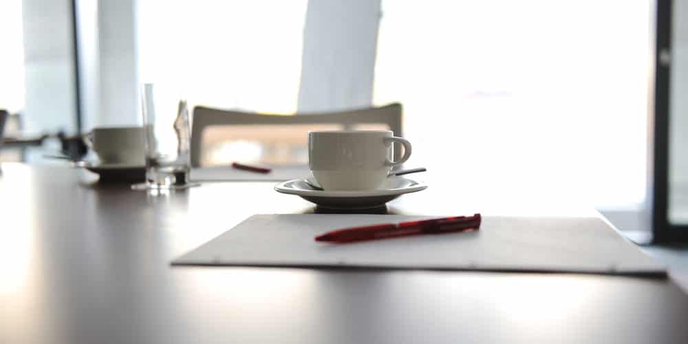 Papeis brancos, caneta vermelha e xícara branca de café sobre mesa de escritório em frente a amplas janelas