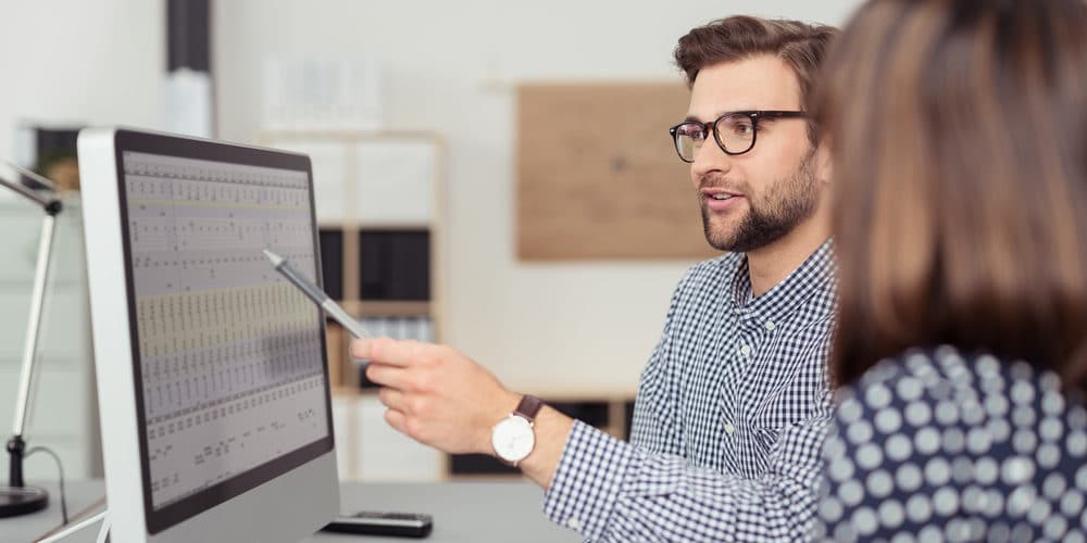 Dupla de trabalhadores em escritório bem iluminado, sentados em frente a computador enquanto o homem explica um plano de negócios para a mulher.
