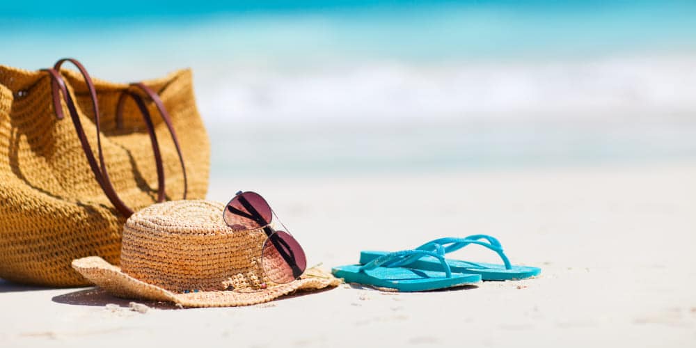 Bolsa de praia, chapéu de palha, chinelos azuis e óculos de sol de lentes rosas sobre areia de praia