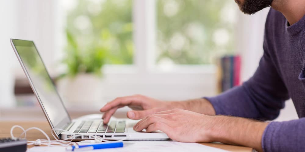 Homem vestindo blusa azul escura de mangas compridas enquanto trabalha de casa em seu laptop prateado