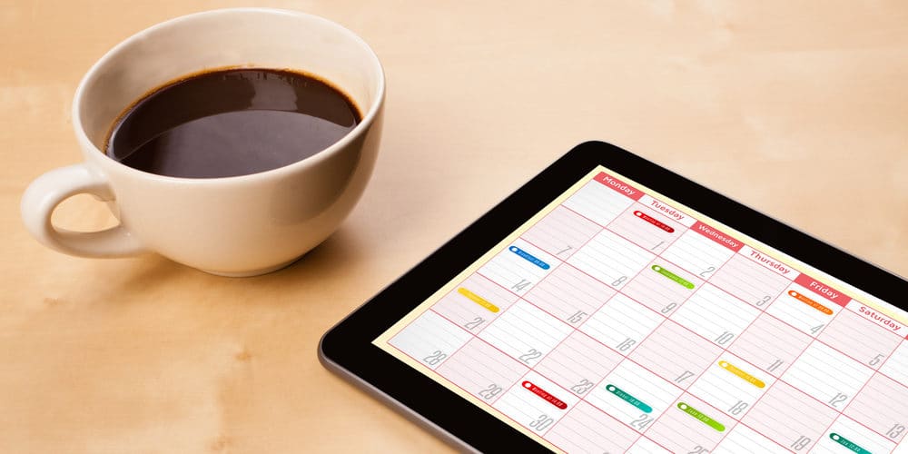 Tablet preto sobre mesa de madeira clara exibindo uma programação em calendário ao lado de uma xícara cheia de café