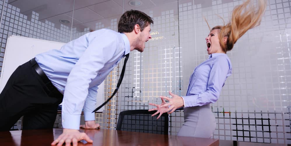 Homem e mulher vestidos com roupa social discutem raivosamente no escritório, dentro de uma sala de reunião
