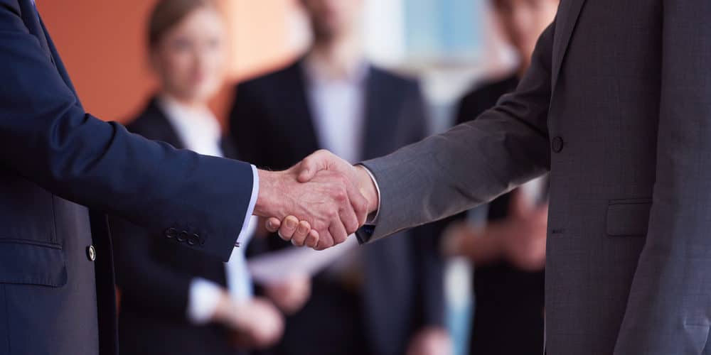 Homens vestidos de terno dando um aperto de mãos em frente a um grupo de três pessoas, fora de foco, ao fundo