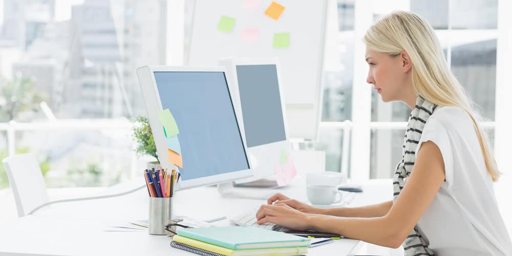 Mulher branca, de cabelos loiros, trabalha em frente ao seu computador em escritório amplo e decoração toda branca