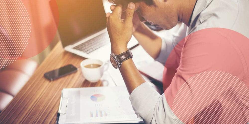 Homem branco sentado à mesa de trabalho com as mãos apoiadas sobre a testa e ao lado de uma xícara de café, um celular e um computador.