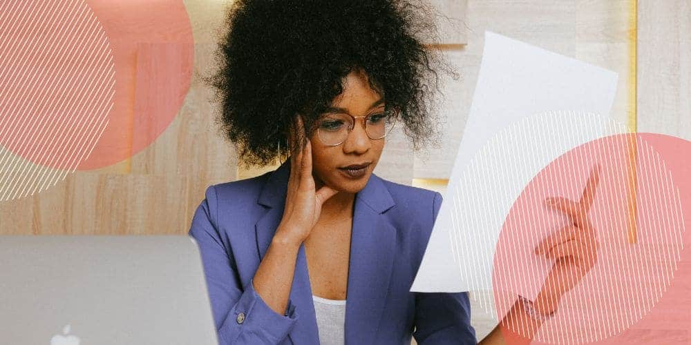 Mulher negra usando um blazer azul escuro e óculos enquanto lê um documento em seu escritório