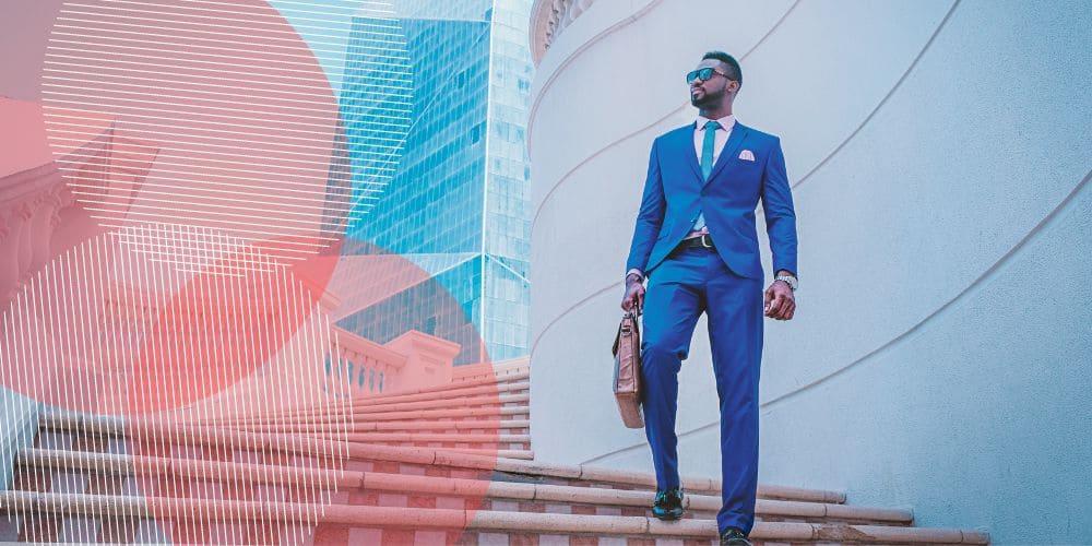 homem negro vestindo um terno azul completo com camisa branca por baixo e óculos escuros. ele segura uma maleta marrom e está descendo as escadas externas de um edifício.