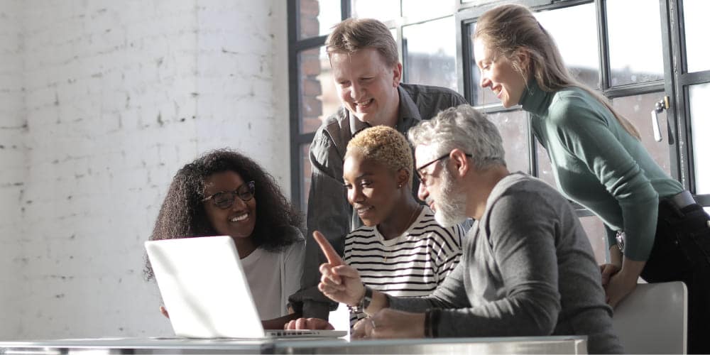 Grupo de cinco colegas reunidos em mesa de trabalho em frente a um laptop