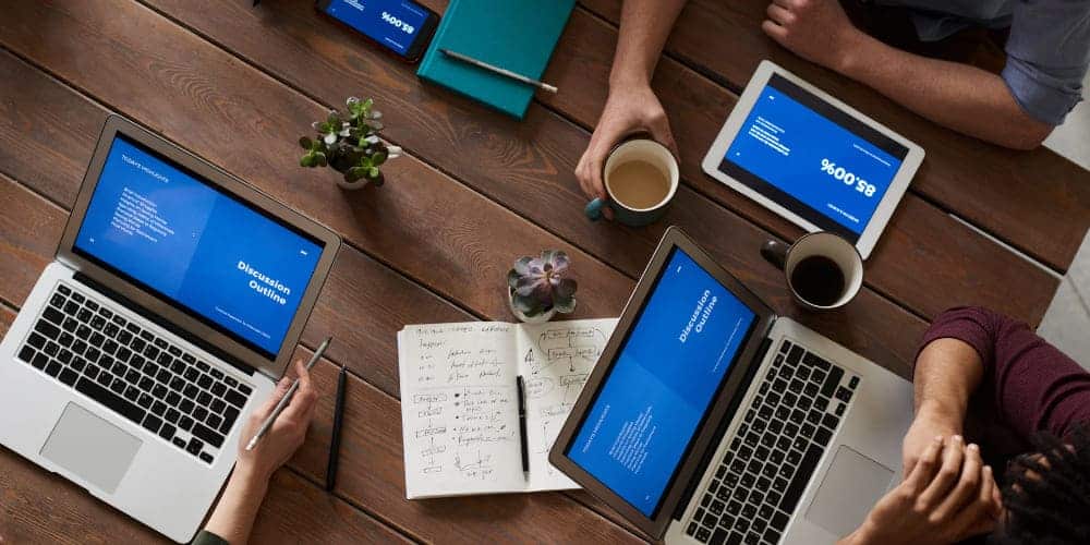 Trabalhadores reunidos ao redor de mesa de trabalho feita de madeira, cada um com seu laptop aberto em uma tela azul