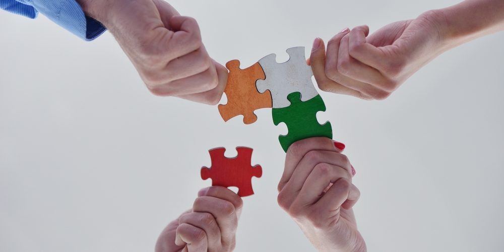 Quatro mãos seguram peças coloridas de quebra-cabeças, nas cores laranja, branco, verde e vermelho, de baixo para cima em frente à câmera