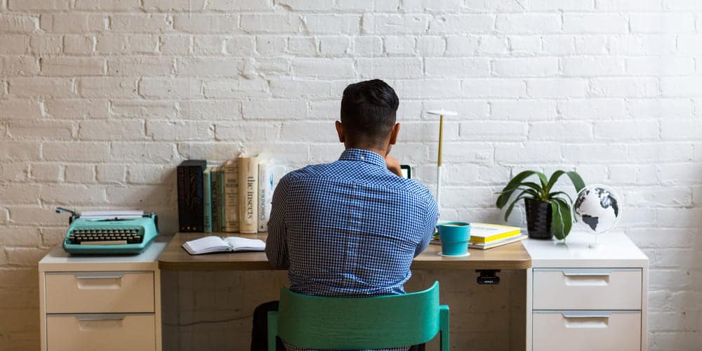 Homem vestido de camisa azul clara está sentado de costas para a câmera enquanto trabalha em sua mesa de escritório, sobre a qual há livros, uma xícara de café e um laptop