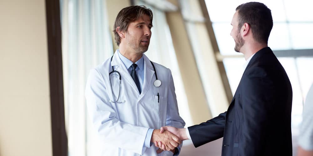 Médico vestido com jaleco e usando um estetoscópio ao redor do pescoço aperta a mão de um homem engravatado após o exame admissional