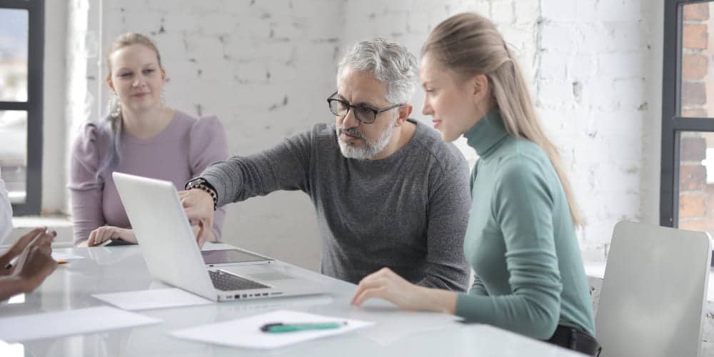 Homem de cabelos brancos e mulher de cabelos loiros sentados em frente a um laptop em mesa de trabalho