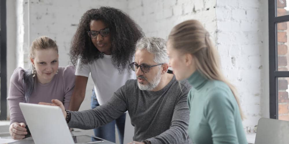 Grupo de quatro colegas de trabalho reunidos ao redor do computador do mais velho deles para avaliar mudanças no plano estratégico