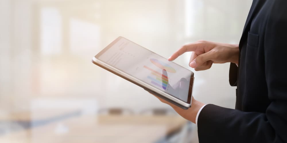 Pessoa vestida com blazer preto segura um tablet com a mão esquerda enquanto clica na tela com o indicador da mão direita