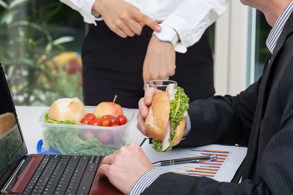 Trabalhador vestido de roupa social almoçando um sanduíche natural em sua mesa de trabalho