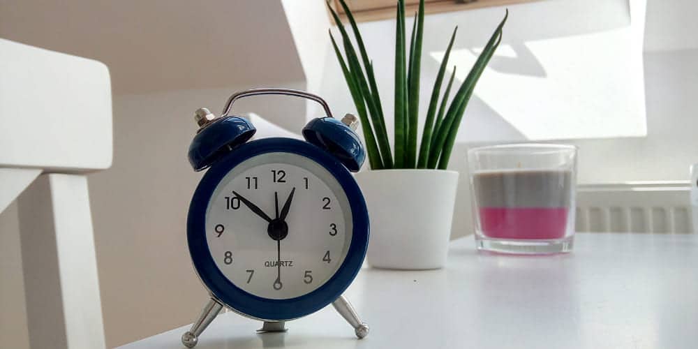 Relógio analógico azul, vaso branco com planta verde e uma vela em pote de vidro sobre uma mesa branca