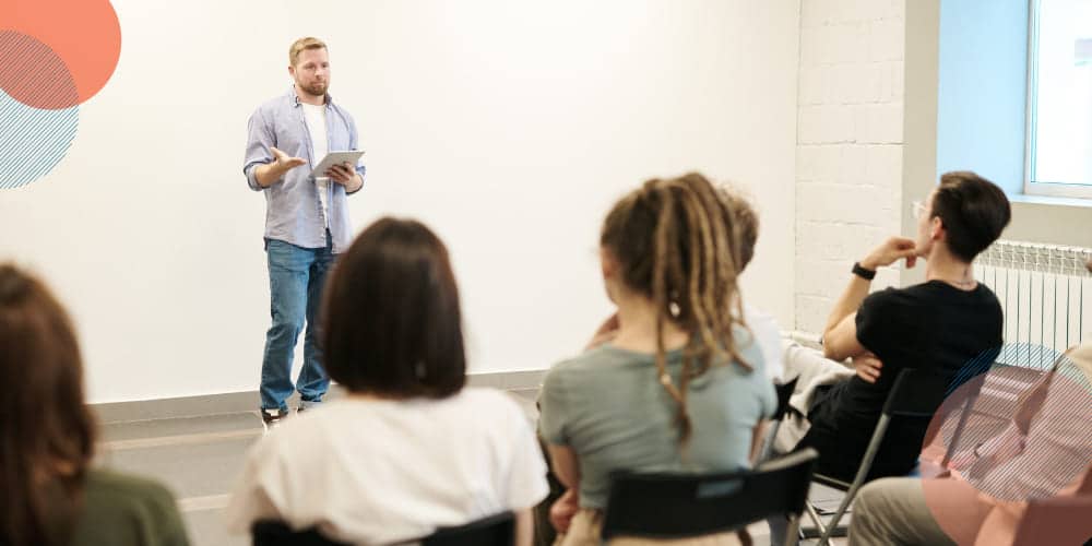 Homem vestido de camisa azul clara e calça jeans fala para plateia sentada em cadeiras pretas de plástico