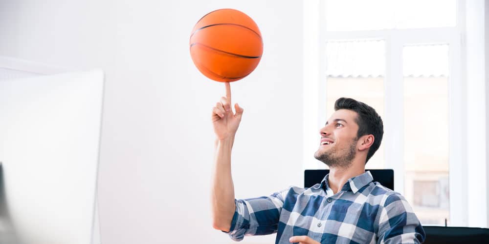 Homem jovem vestindo camisa xadrez azul e branca está sentado em uma cadeira de escritório, em frente a uma mesa, enquanto gira uma bola de basquete laranja em seu dedo indicador