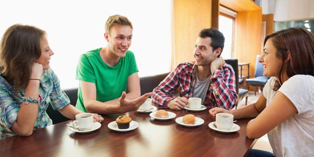 Grupo de amigos vestidos informalmente tomando café e comendo um lanche da tarde em mesa de madeira escura.