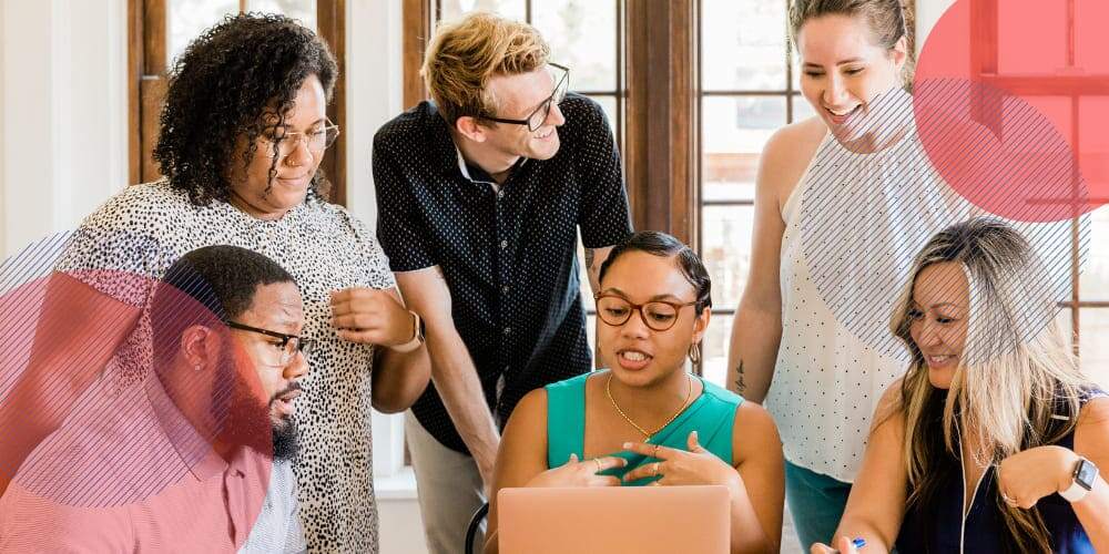 Equipe de trabalho ao redor de mesa em escritório na qual há uma profissional mostrando informações em seu laptop