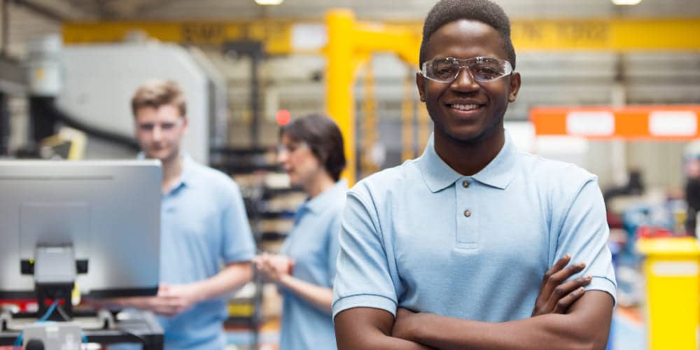 Jovem aprendiz vestindo camisa polo azul-claro e óculos de proteção enquanto sorri para a foto em ambiente de fábrica